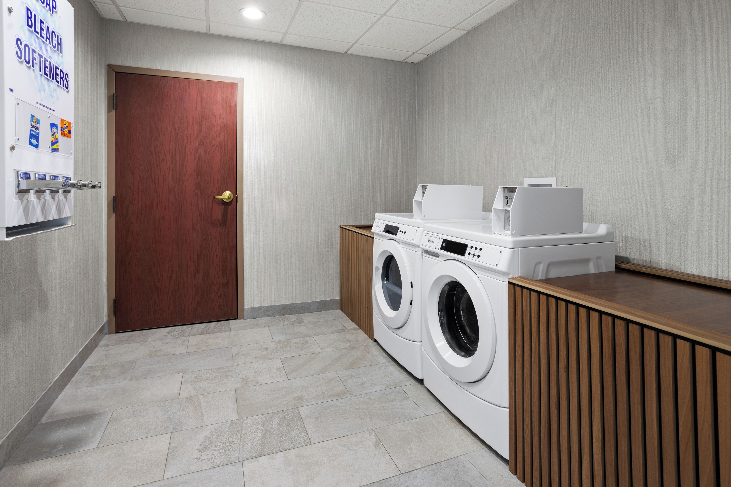 Un cuarto de lavandería con puerta de madera, lavadora, secadora y dispensador de detergente.
