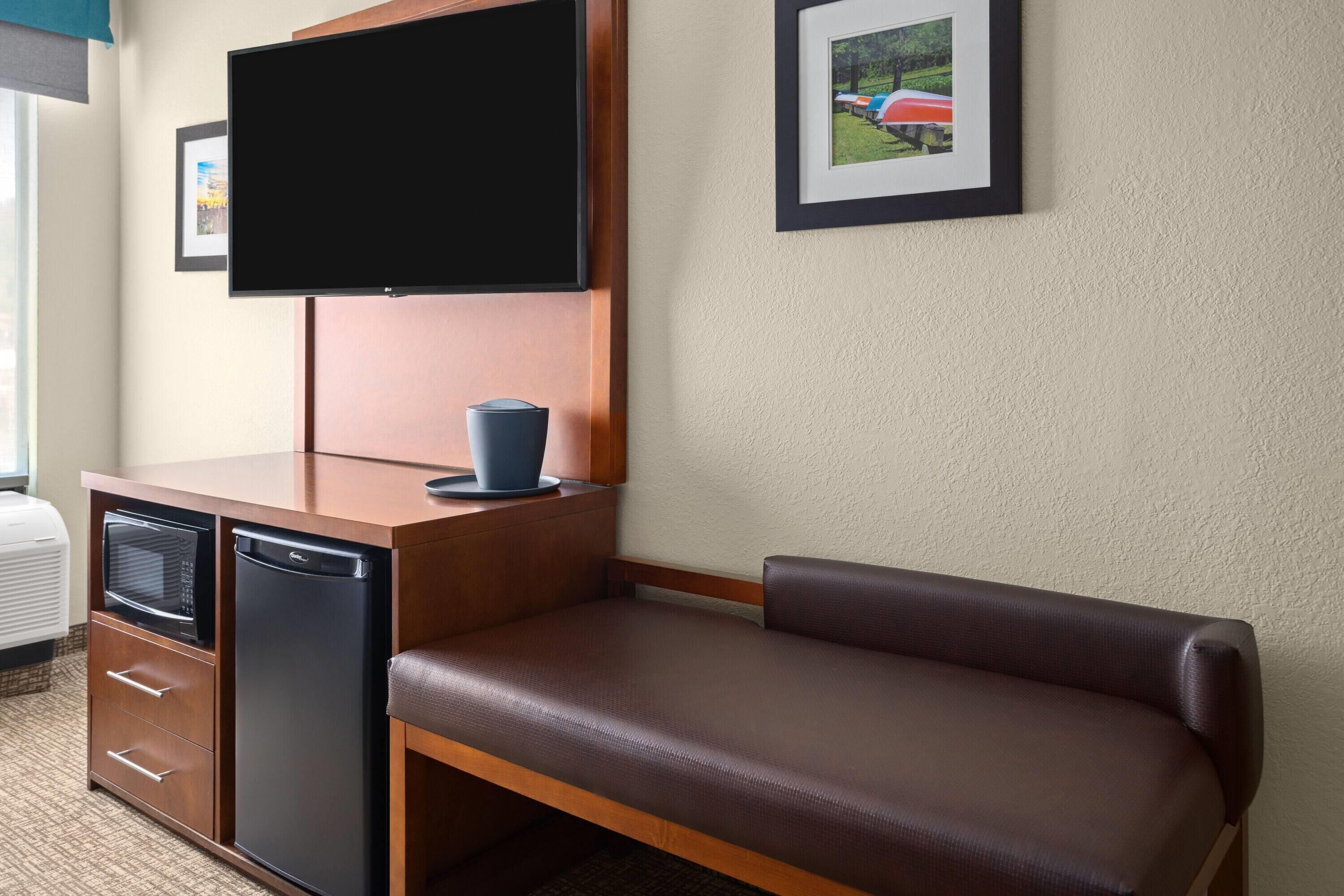 Quarto de hotel com televisão de tela plana na parede, pequena cozinha compacta com micro-ondas e geladeira, um banco de couro marrom e um quadro pendurado na parede.