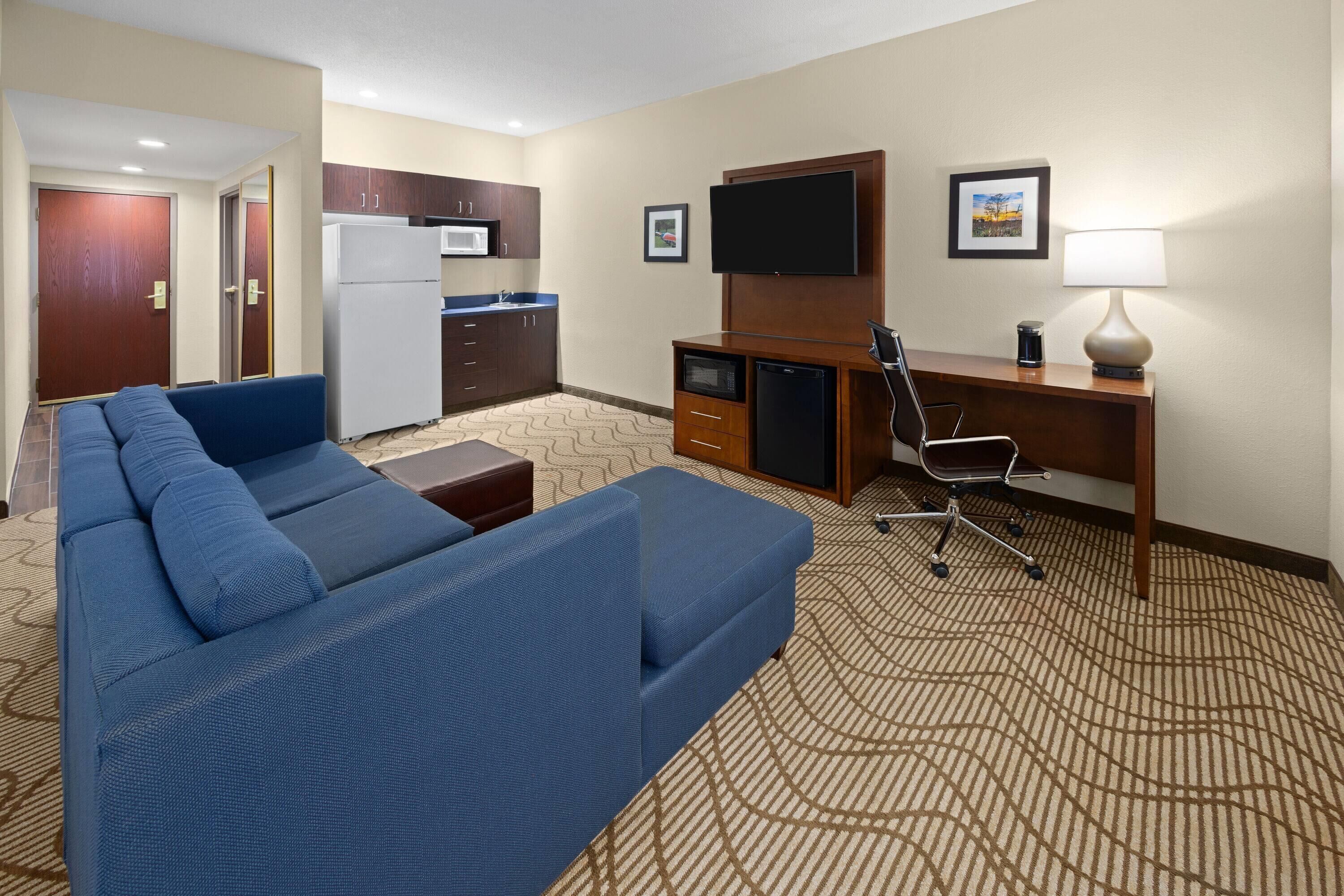 A hotel room with a blue sofa, a desk with a computer, a TV, a microwave, and a refrigerator.
