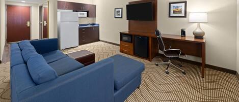 A hotel room with a blue sofa, a desk with a computer, a TV, a microwave, and a refrigerator.
