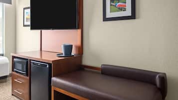 A hotel room with a flat-screen TV mounted on the wall, a small kitchenette with a microwave and refrigerator, a brown leather sofa, and a framed picture on the wall.