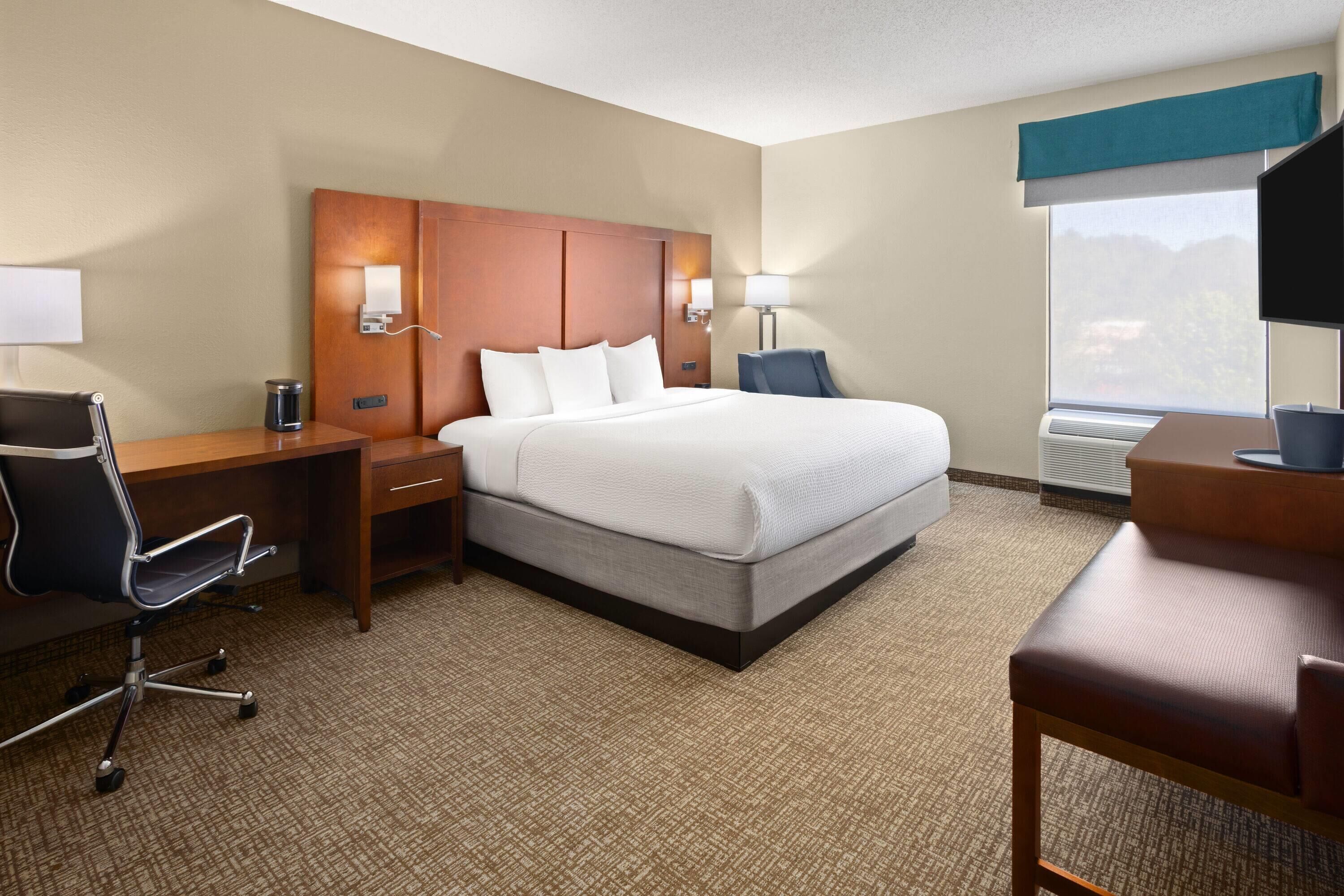 A hotel room with a large bed, a desk with a chair, a desk lamp, a nightstand, and a window with a view of the outdoors.