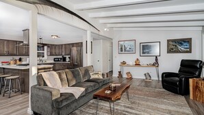A modern living room with a grey sofa, a wooden coffee table, and a black leather chair. There is a kitchen in the background with wooden cabinets and stainless steel appliances. - Private Home on the Kenai River at Gull's Landing (Soldotna)