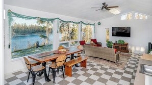 A spacious living area with a large window overlooking a lake, a dining table with chairs, a sofa, and a television mounted on the wall. - Private Home on the Kenai River at Gull's Landing (Soldotna)