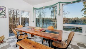 A dining area with a large wooden table and chairs, a checkered floor, and a view of a lake and trees through large windows. - Private Home on the Kenai River at Gull's Landing (Soldotna)