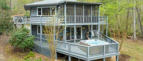 Eine zweistöckige Holzhütte mit Whirlpool auf der Terrasse, umgeben von Bäumen.
