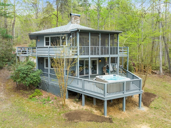 A two-story cabin with a hot tub on the deck, surrounded by trees.