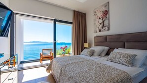 Une chambre avec un grand lit, une vue sur la mer, un fauteuil près de la fenêtre et un tableau au mur.