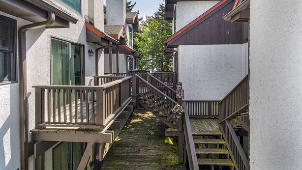 A narrow wooden staircase leading to a landing with a railing, flanked by buildings with white walls and dark roofs. - Whistlepunk 36: 1 Br Plus Bonus Sleeping Loft Slope Side (Snowshoe)
