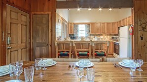 A wooden dining table set for six with chairs, a kitchen in the background, and wooden paneling. - Whistlepunk 13 Spacious 4 Br Condo Slope Side (Snowshoe)