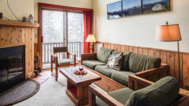 A cozy living room with a fireplace, wooden furniture, a dining table with wine glasses, and a view of mountains through the window.