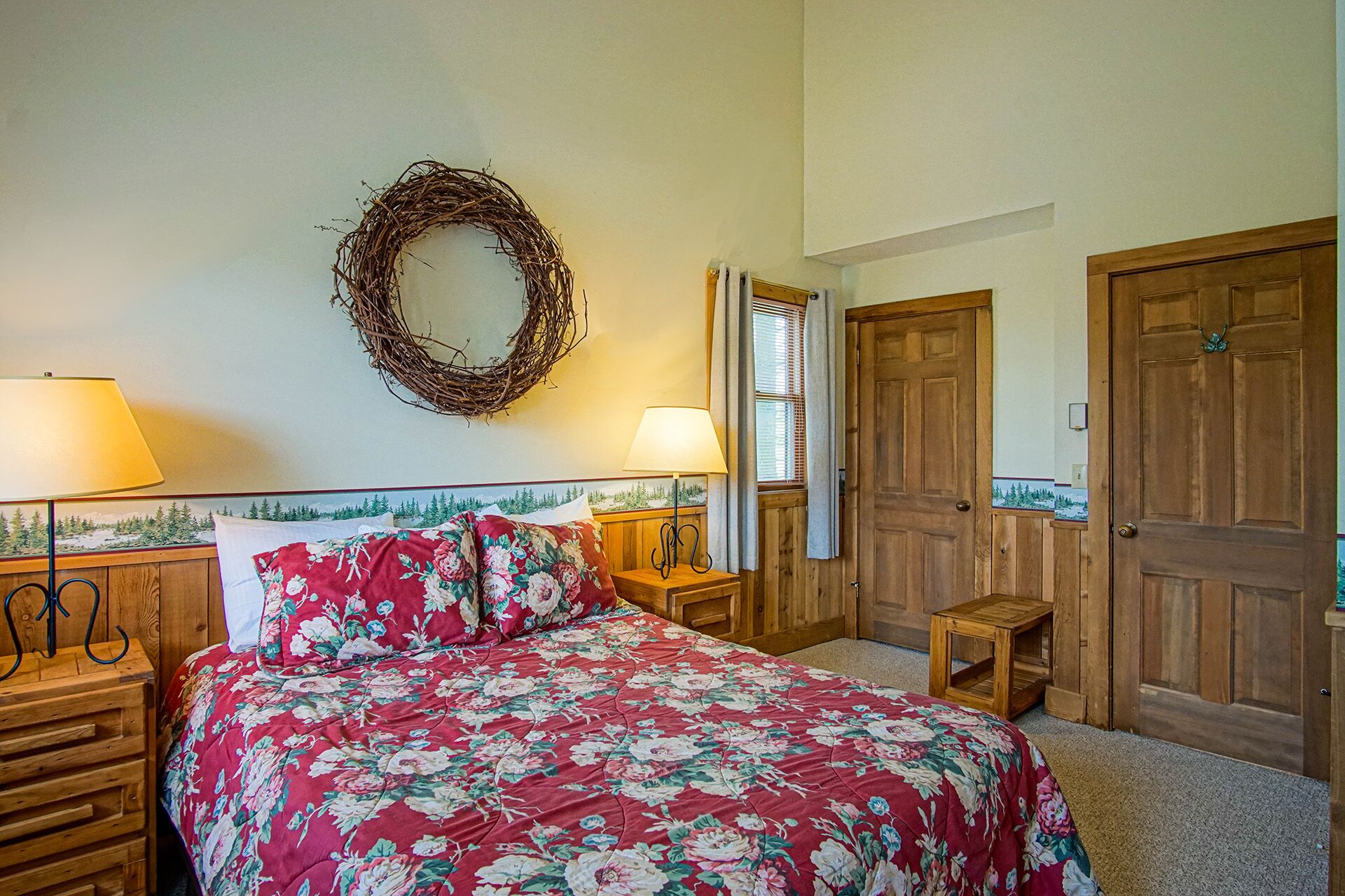 Ein Schlafzimmer mit geblümter Bettdecke, einem Holzkopfteil, einem an der Wand befestigten Kranz, zwei Nachttischlampen und einer Tür mit Spiegel.