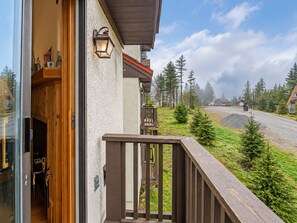 A wooden deck with a railing, a glass door leading to a room, and a view of a gravel road with trees and houses in the background.
