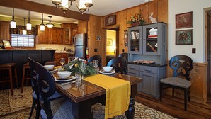 Ein Essbereich mit Holztisch, Stühlen und einer Kronleuchter. Im Hintergrund ist eine Küche mit Holzausbau und einem Kühlschrank zu sehen.