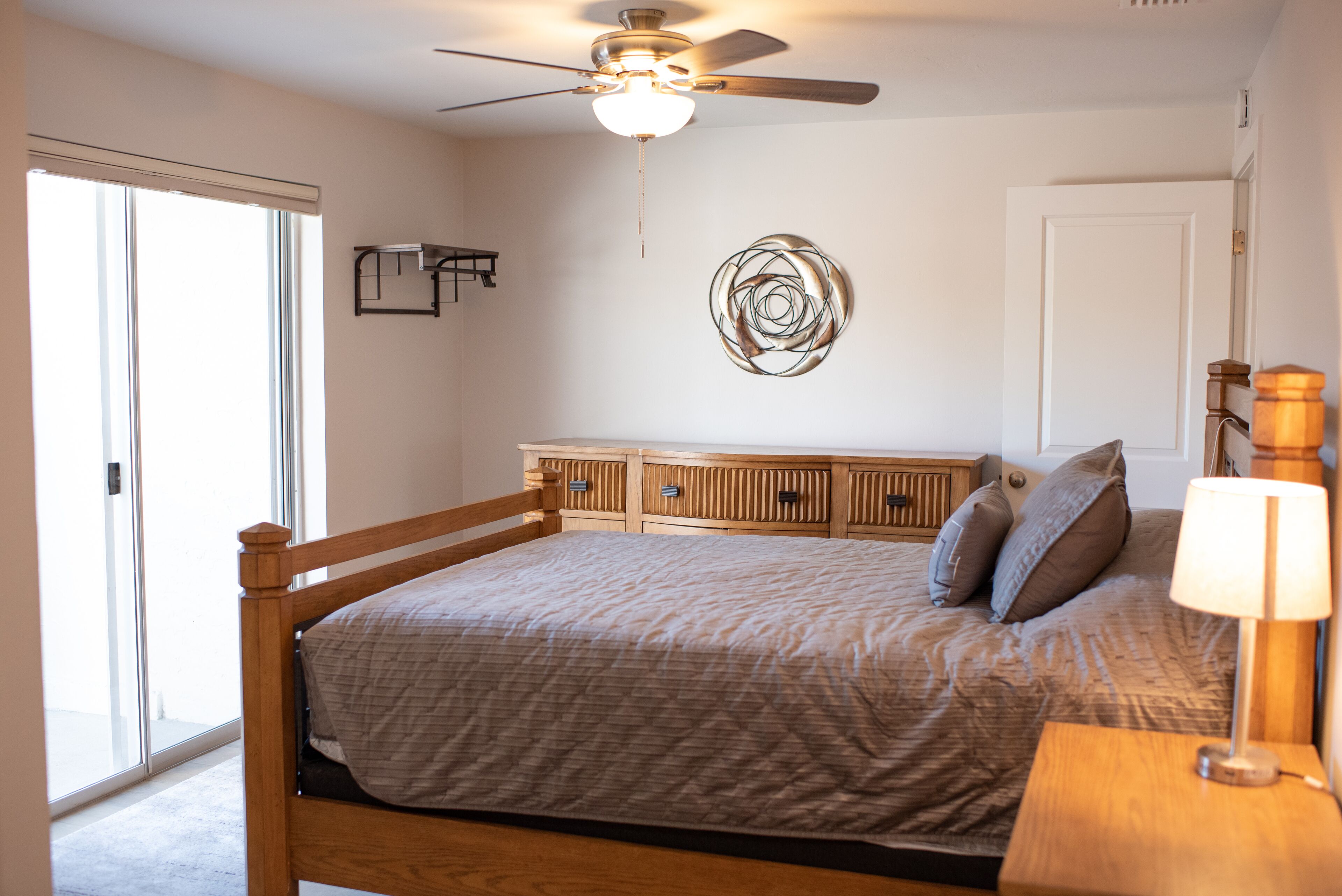 A bedroom with a bed, a ceiling fan, a wall-mounted light, a door, and a window.