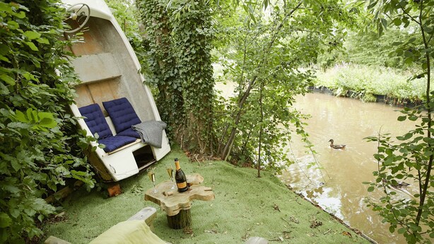 A wooden boat with blue cushions and a bottle of champagne on a grassy area beside a river. - Two bedroomed property situation in a beautiful secluded location with patio area overlooking fields (Wootton Wawen)