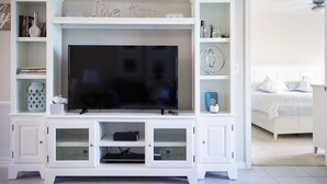 A living room with a flat-screen TV on a white entertainment unit, a bookshelf, and a bed in the background. - Vacation in Paradise. New Kitchen, New bedroom furniture. (Naples)
