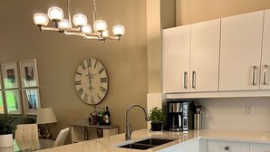 A modern kitchen with white cabinetry, a central island with a dishwasher, a sink, and a coffee maker. There is a large wall clock and framed pictures on the wall. - Vacation in Paradise. New Kitchen, New bedroom furniture. (Naples)