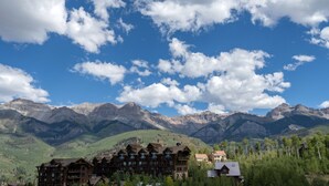 A lodge with multiple wooden buildings, surrounded by mountains and trees under a partly cloudy sky.