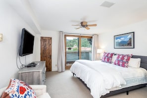 Une chambre à coucher comprenant un lit, un téléviseur, un ventilateur de plafond, une fenêtre avec des rideaux et un tableau encadré accroché au mur.