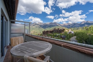 A balcony with a wooden table and chairs, overlooking a mountainous landscape with a building complex in the distance.