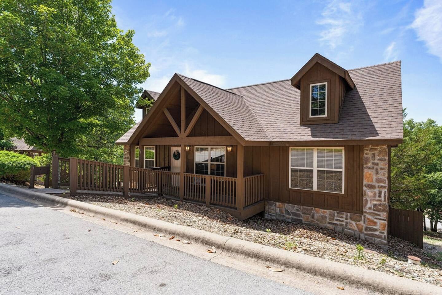 A two-story wooden house with a stone foundation, a porch with wooden railings, and a fenced driveway.