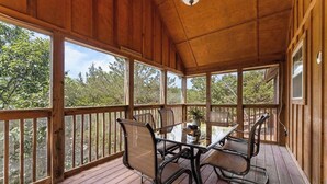 Una terraza cubierta al aire libre con una mesa de vidrio y sillas de metal, rodeada de barandas de madera y un piso de madera.