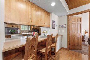 A kitchen with wooden cabinets, a dining table with chairs, and a view into a bedroom through an open door. - Sunshine Stonebridge Cabin near Silver Dollar City - Pools, Lake, Golf! (Branson West)