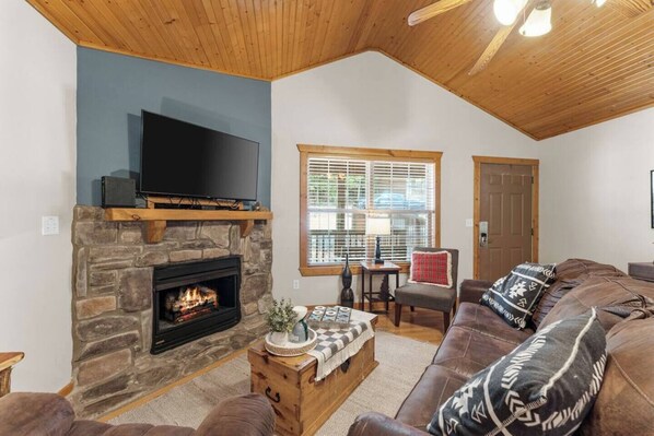 A cozy living room with a stone fireplace, a flat-screen TV mounted above it, a comfortable couch with patterned cushions, and a wooden coffee table. - Sunshine Stonebridge Cabin near Silver Dollar City - Pools, Lake, Golf! (Branson West)