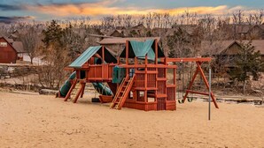 Una estructura de juego de madera con toboganes y columpios sobre una playa arenosa, rodeada de casas y árboles.
