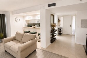 Una sala de estar con un sofá color beige, una zona de comedor con una cocina al fondo y un pasillo que lleva a una puerta.