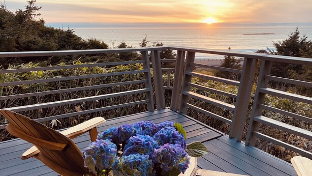 A wooden deck with a railing, a wooden chair, a table with a vase of flowers, and a view of the ocean at sunset. - Breathtaking ocean views & adventure at your doorstep (Newport)