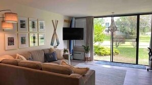 A living room with a large sofa, a flat-screen TV mounted on the wall, and a sliding glass door leading to a garden. - Hidden Gem! Turtle Bay Hideaway (Kahuku)