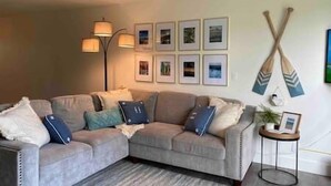 A living room with a grey sectional sofa, a floor lamp, framed pictures on the wall, and two paddleboards hanging on the wall. - Hidden Gem! Turtle Bay Hideaway (Kahuku)