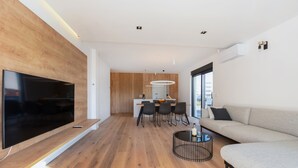 A modern living room with a flat-screen TV, a sofa, a dining area with chairs, and a kitchen in the background.