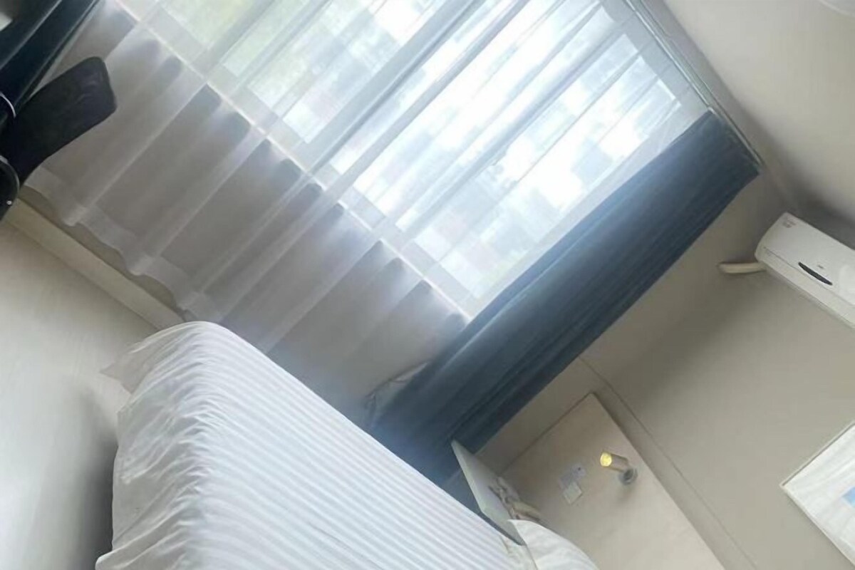 Ett hotellrum med en säng, ett fönster med gardiner, en luftkonditioneringsanläggning och en väggmonterad lampa.