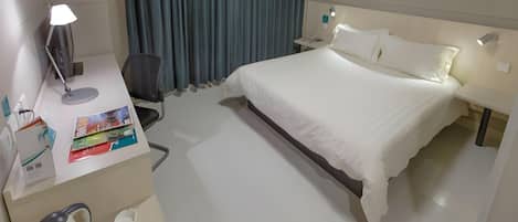 A hotel room with a bed, desk, chair, lamp, and a painting on the wall.