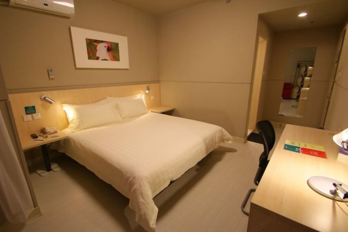 A hotel room with a bed, bedside table, lamp, desk, chair, and a framed picture on the wall.