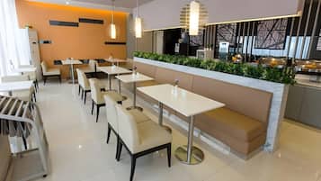 Interior de un restaurante moderno con mesas y bancos blancos, iluminación colgante y una barra con un arreglo de plantas verdes.