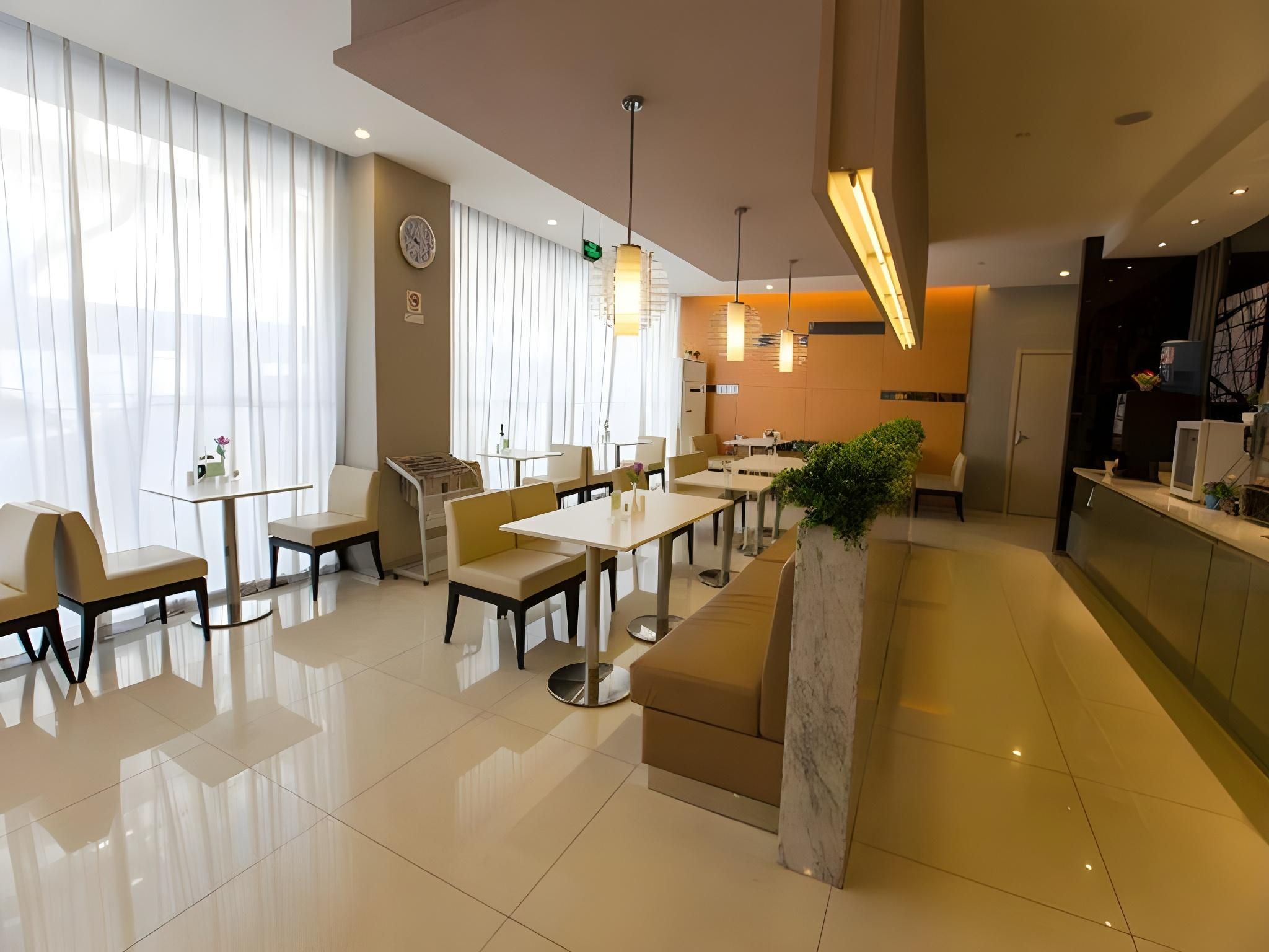 Un comedor moderno de hotel con varias mesas y sillas, un mostrador de recepción y un gran ventanal con cortinas.