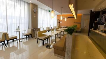 Un comedor moderno de hotel con varias mesas y sillas, un mostrador de recepción y un gran ventanal con cortinas.