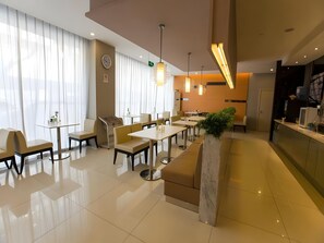 Un comedor moderno de hotel con varias mesas y sillas, un mostrador de recepción y un gran ventanal con cortinas.