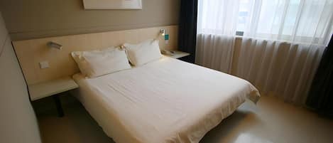 A hotel room with a large bed, a bedside table with a lamp, a painting on the wall, and a window with curtains.