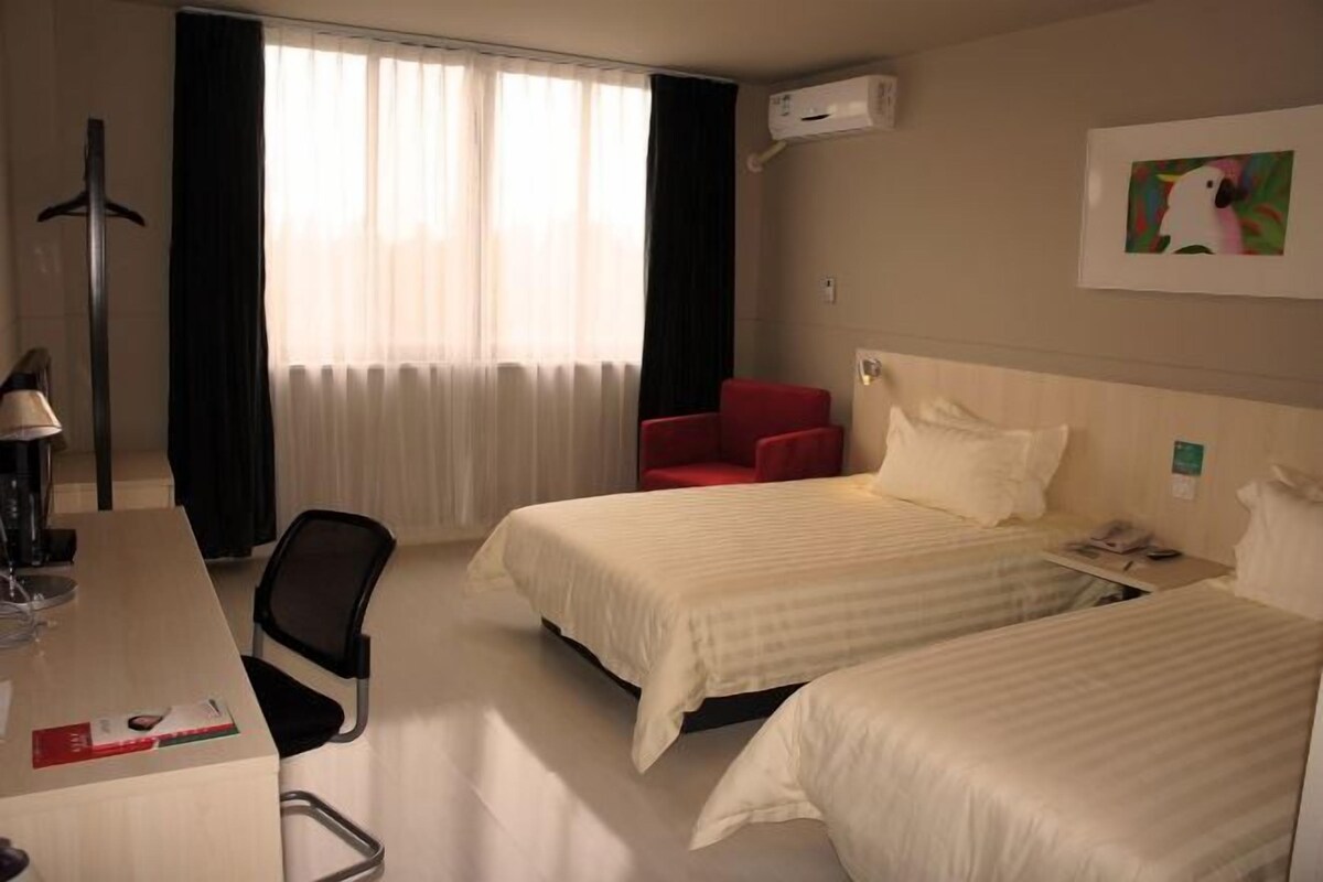 A hotel room with two beds, a desk, a chair, a red armchair, a window with curtains, an air conditioning unit, and a framed picture on the wall.