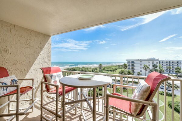 A balcony with a table and chairs, overlooking a beach and a building. - Shoreline view for miles- Oceanfront heated pool (Cape Canaveral)