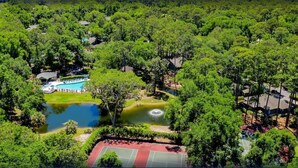 Eine Vogelperspektive zeigt ein Resort mit Swimmingpool, Teich und Tennisplätzen, umgeben von üppiger Vegetation.