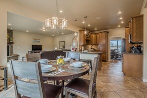 A spacious dining area with a round table set for six, surrounded by wooden chairs. A chandelier hangs above, and a stone fireplace is visible in the background. - Sunshine II – Bike Path Access, Close to Town (Fruita)
