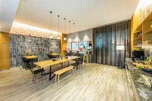 A modern dining area with a long wooden table, black chairs, and a bench. There are pendant lights above the table, and a bar area with stools is visible in the background. - Sofis Pop Hotel (Fuzhou)