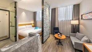 A hotel room with a bed, a sofa, a small table, a chair, a desk, a lamp, a painting, and a shower area. - Sofis POP Hotel (Fuzhou)