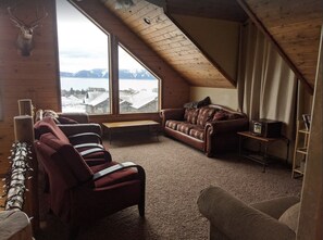 A cozy loft-style room with a large window offering a view of a snowy landscape. There is a leather sofa, a wooden bench, and a small table with a radio.